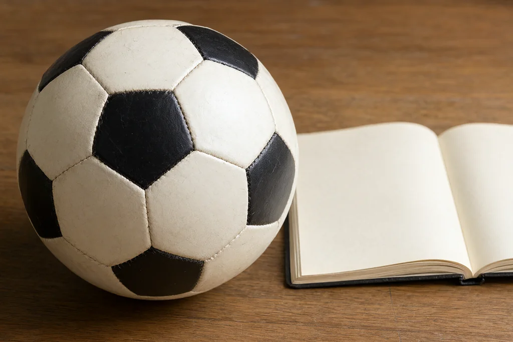 Classic round stitched football with black pentagon panels on a wooden desk beside a blank notebook
